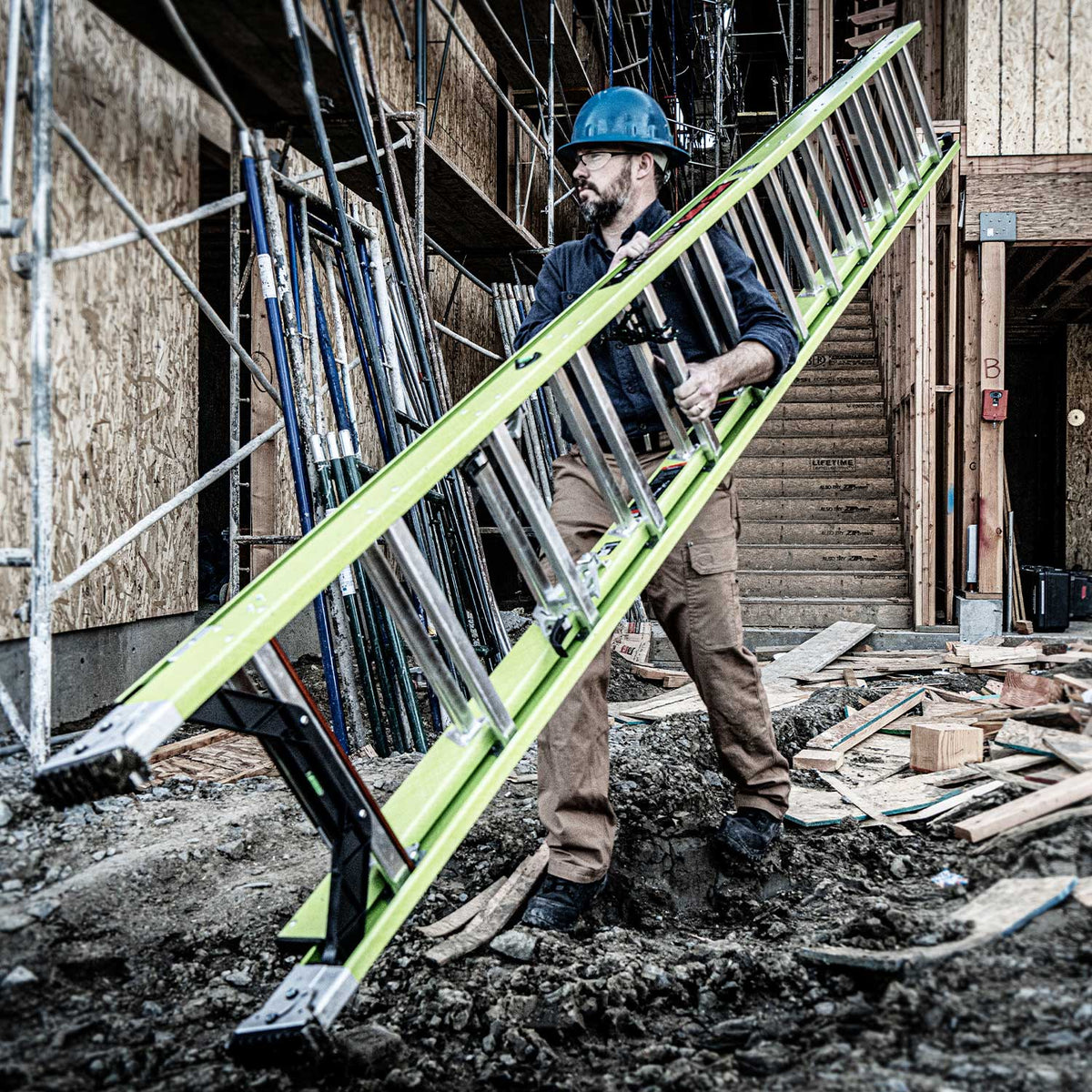 Construction worker carrying Hyperlite Extension ladder across uneven ground at construction site
