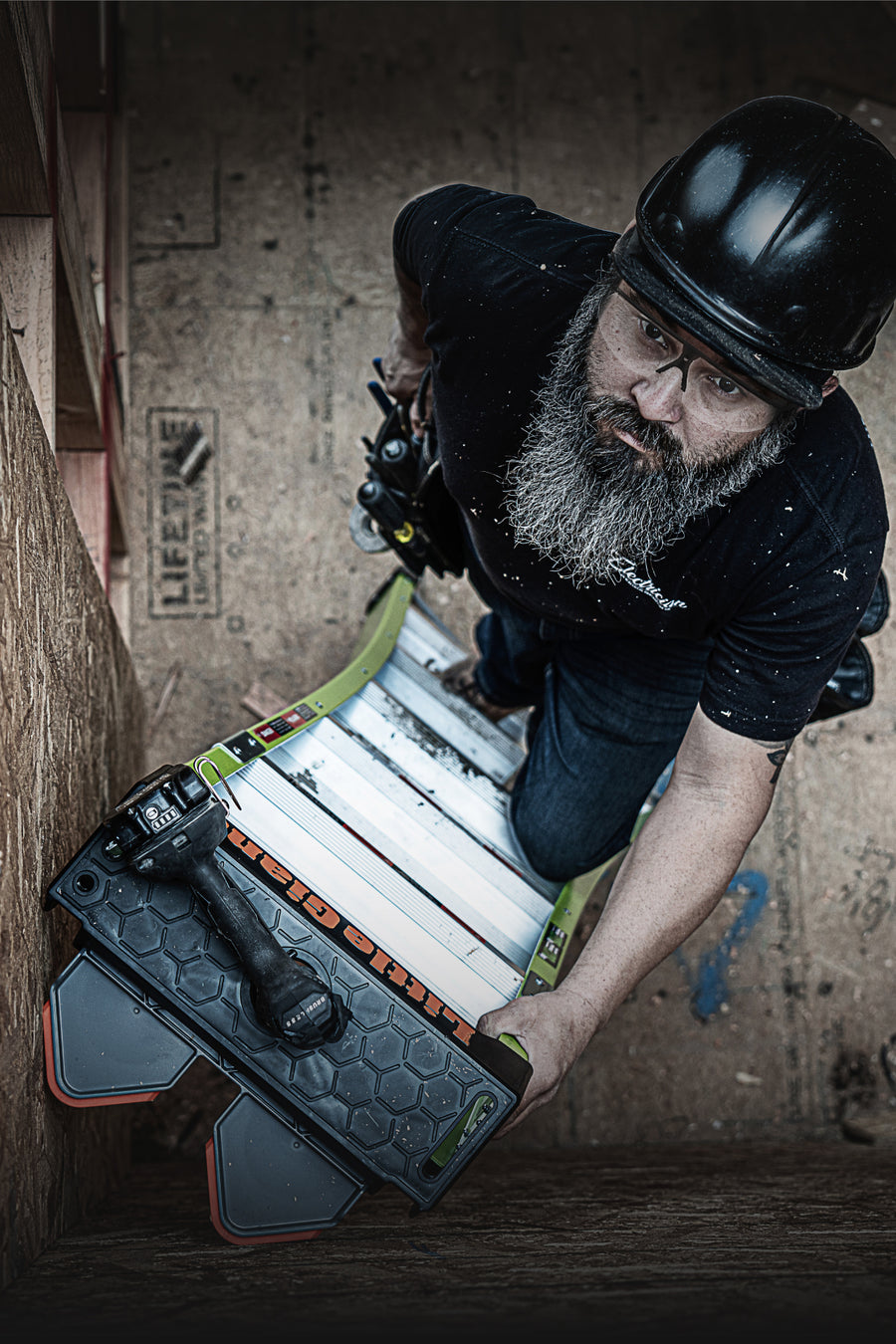 A bearded man wearing a black helmet and glasses kneels on a King Kombo ladder holding a black and green decked board with a drill resting on top. The background is a rustic, textured wooden wall.