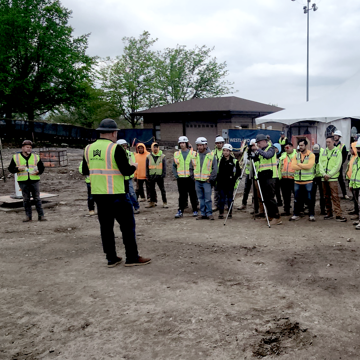 Group of people in safety vests and hard hats standing on a construction site with a speaker addressing them.